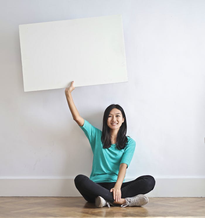 creative-02 Cheerful Asian woman sitting cross legged on floor against white wall in empty apartment and showing white blank banner