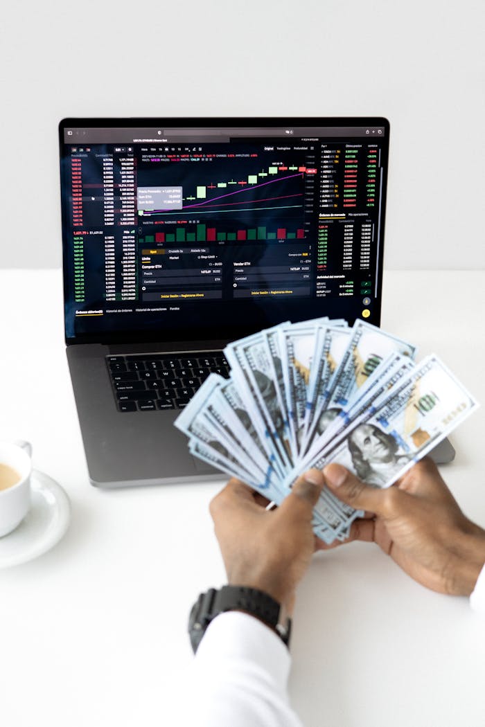 mobile-01 Hands holding cash in front of a laptop displaying stock market data, symbolizing financial growth and investment.