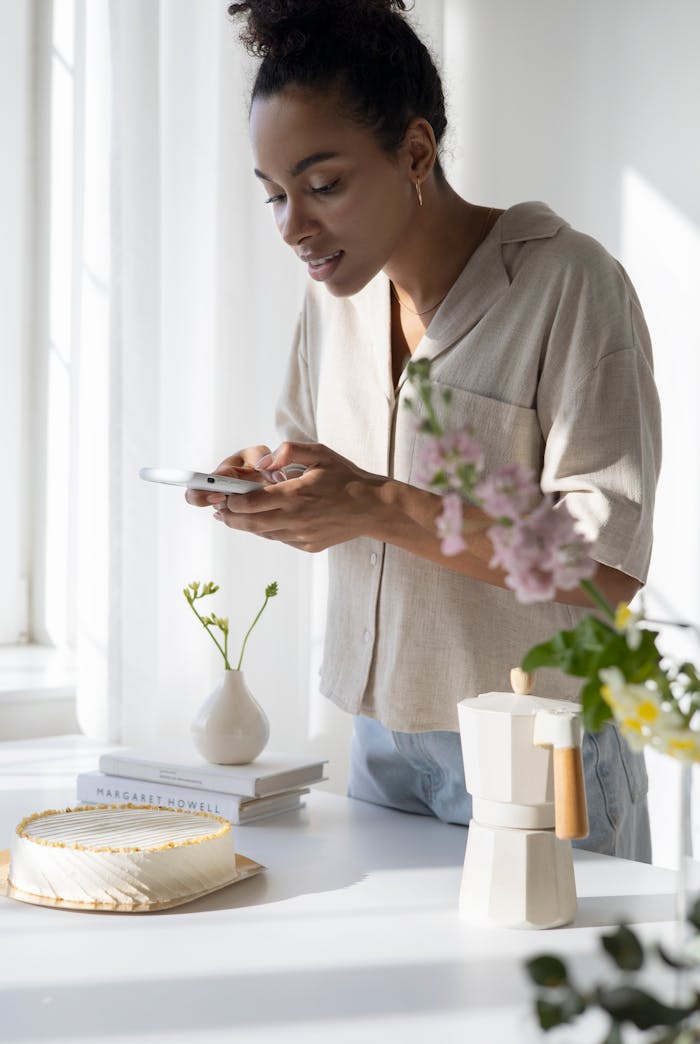 Woman capturing a stylish coffee setup in a sunlit kitchen with flowers and cake.